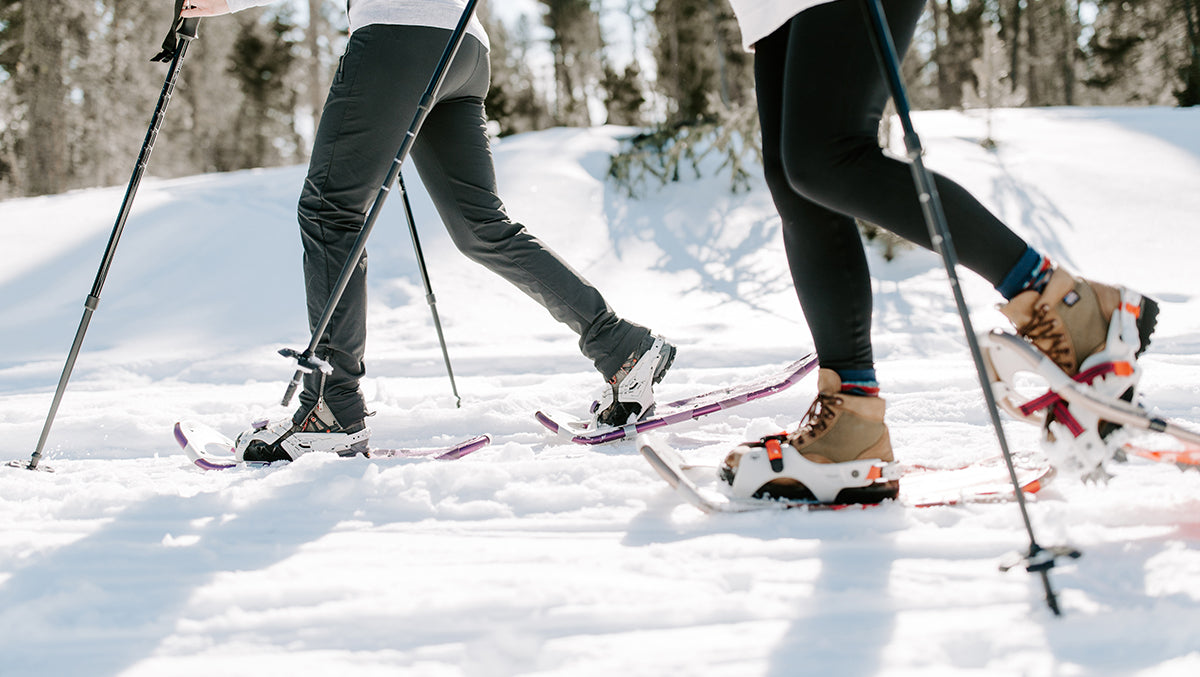 Two women snowshoeing on a trail. Mad Dog's Ski and Board carries a variety of snowshoes, snowshoe/hiking poles and snowshoe accessories for all your winter adventure needs!